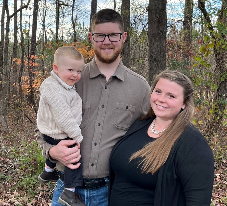 Hidden+Acres+Farm+NC+-+7DF25BEB-2A84-48BD-A573-C571E8E28EF4+(2) (1) Family portrait in a wooded area, featuring a smiling couple with a toddler. The father holds the child, who is wearing a light sweater, while the mother stands beside them, dressed in a black top and pearl necklace. Autumn foliage is visible in the background.