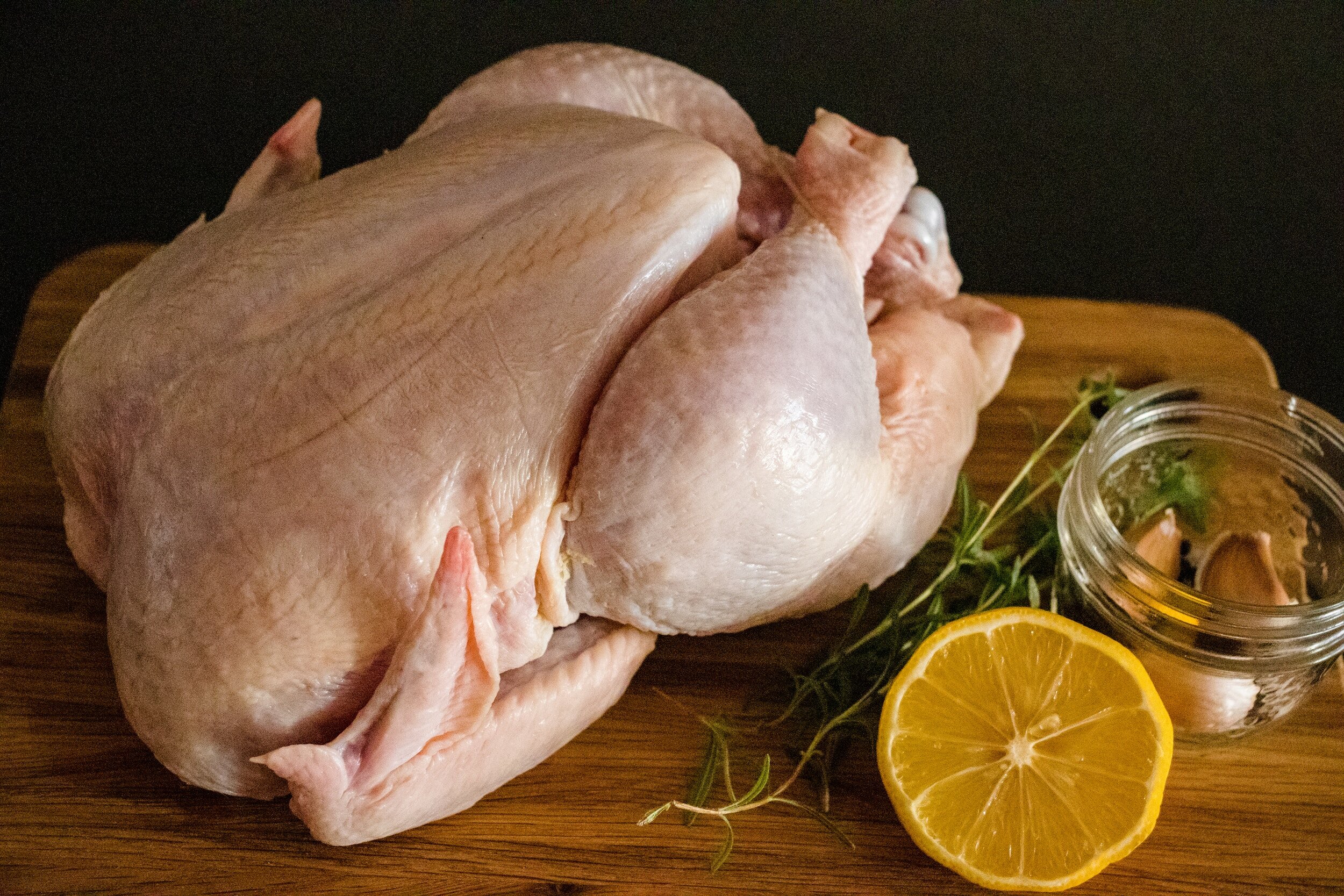 Raw whole chicken on a wooden cutting board, accompanied by fresh herbs, a sliced lemon, and a small jar containing garlic cloves, ideal for meal preparation and cooking recipes.