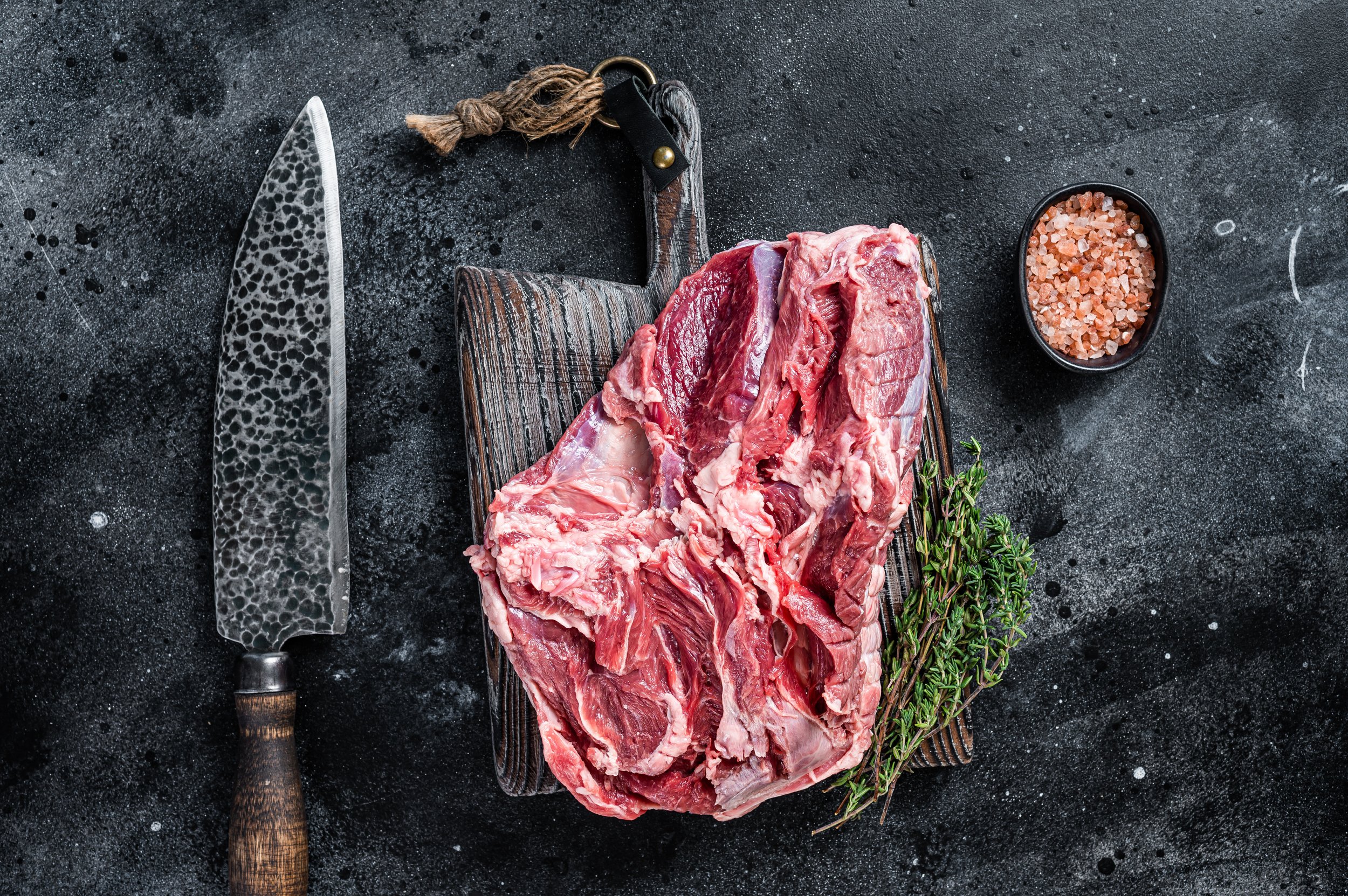 Raw beef cut on a wooden cutting board, accompanied by fresh thyme and a small bowl of pink Himalayan salt, with a traditional Japanese chef's knife nearby, set against a dark textured background.