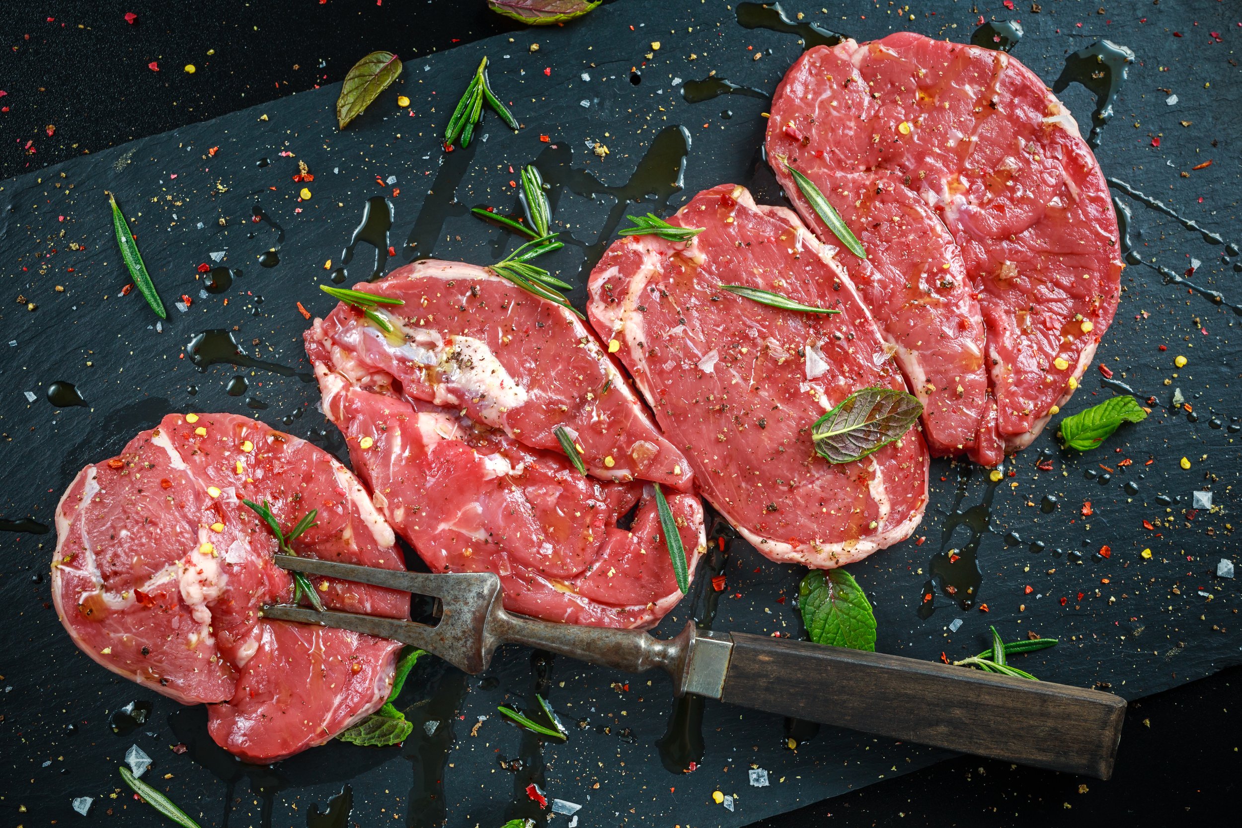Freshly cut beef steaks seasoned with herbs and spices on a black slate board, garnished with rosemary and mint leaves, perfect for grilling or cooking.