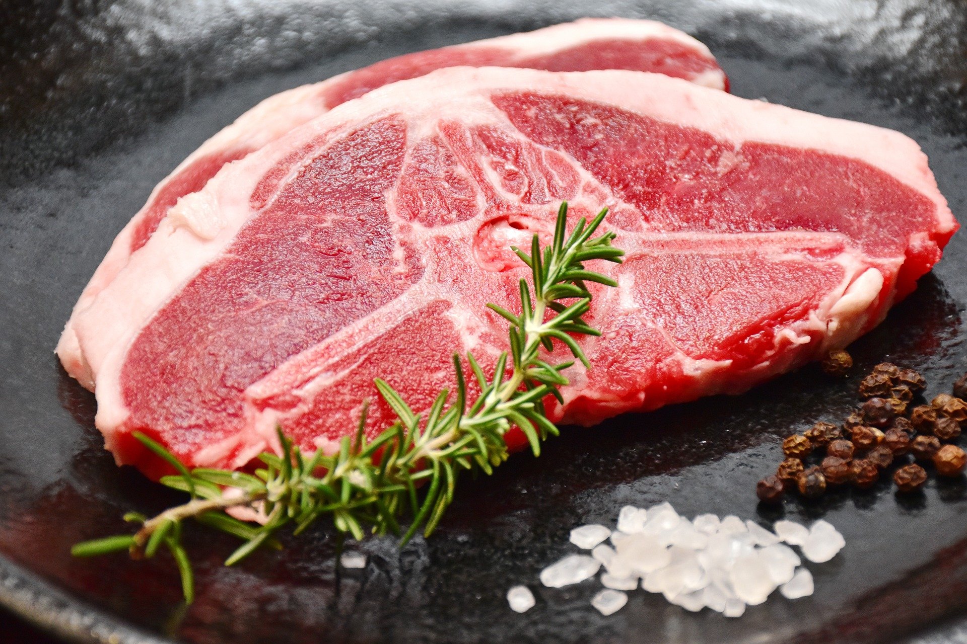 Fresh uncooked lamb chops on a black plate, garnished with rosemary and surrounded by coarse sea salt and black peppercorns, ideal for a gourmet meal.