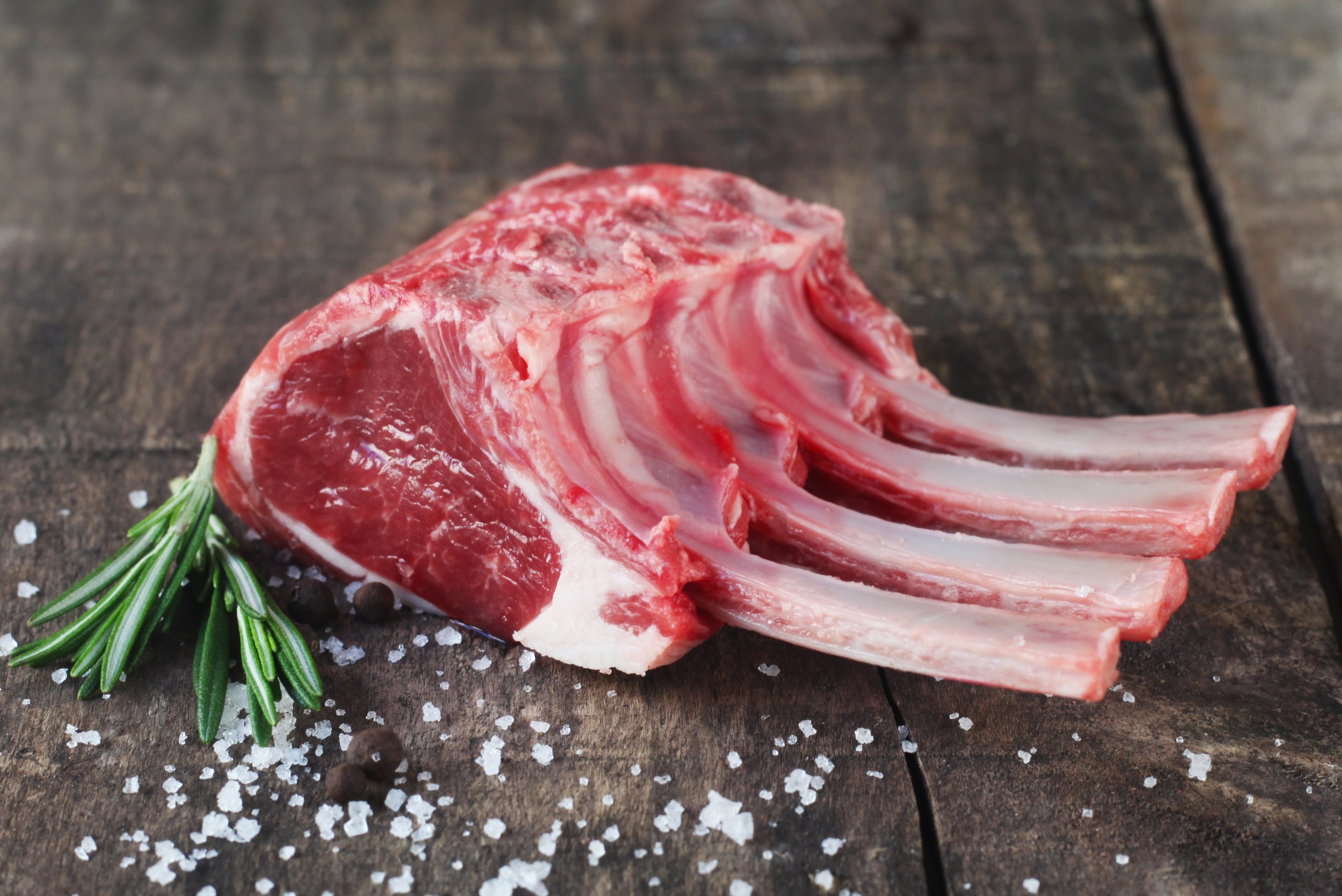 Raw lamb rack with four ribs, garnished with fresh rosemary and coarse sea salt, placed on a rustic wooden surface. Perfect for gourmet cooking and grilling.