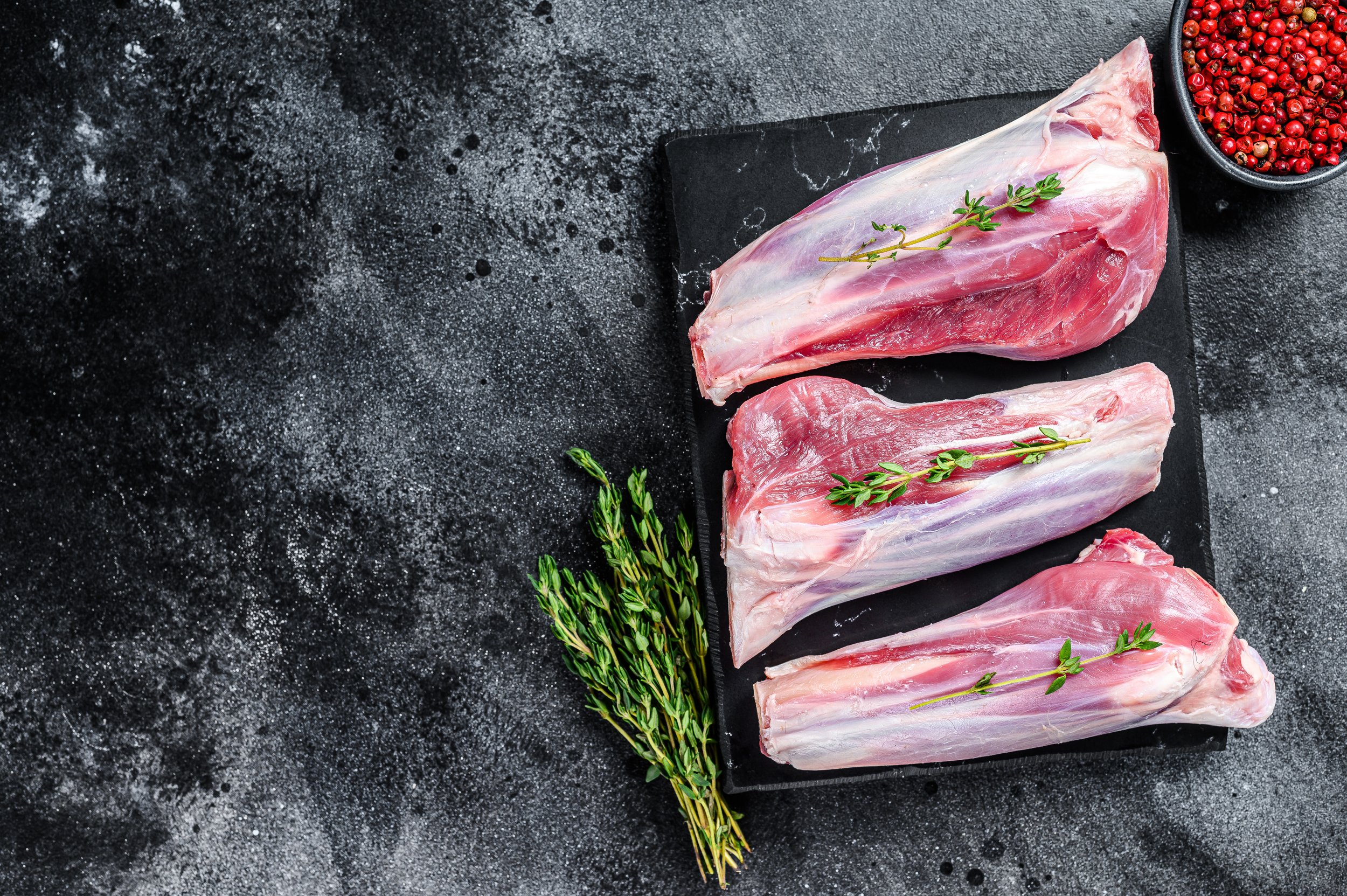 Raw meat cuts on a black stone surface, garnished with fresh thyme, accompanied by a bowl of pink peppercorns, ideal for culinary presentations and gourmet recipes.