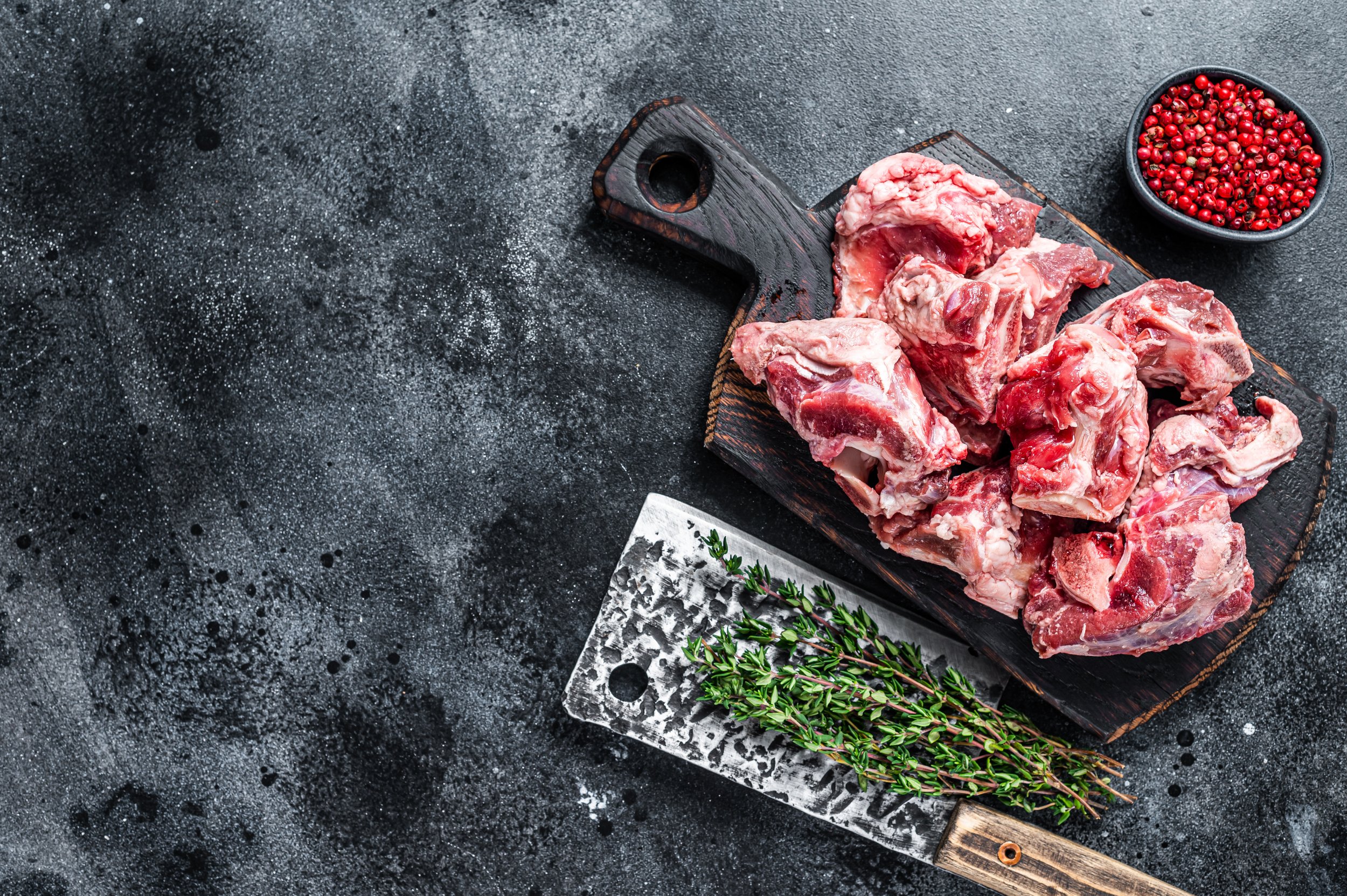 Fresh raw meat pieces on a wooden cutting board alongside a chopping knife and sprigs of thyme, with a bowl of pink peppercorns in the background, ideal for culinary preparation and recipes.