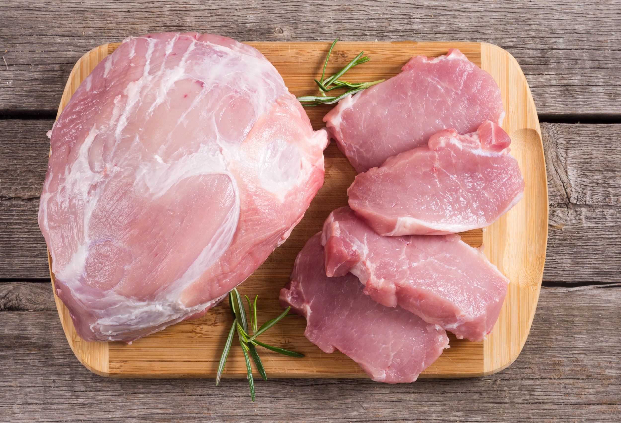 Fresh pork cuts displayed on a wooden cutting board, featuring a large pork shoulder and several pork chops, garnished with sprigs of rosemary for a rustic presentation.