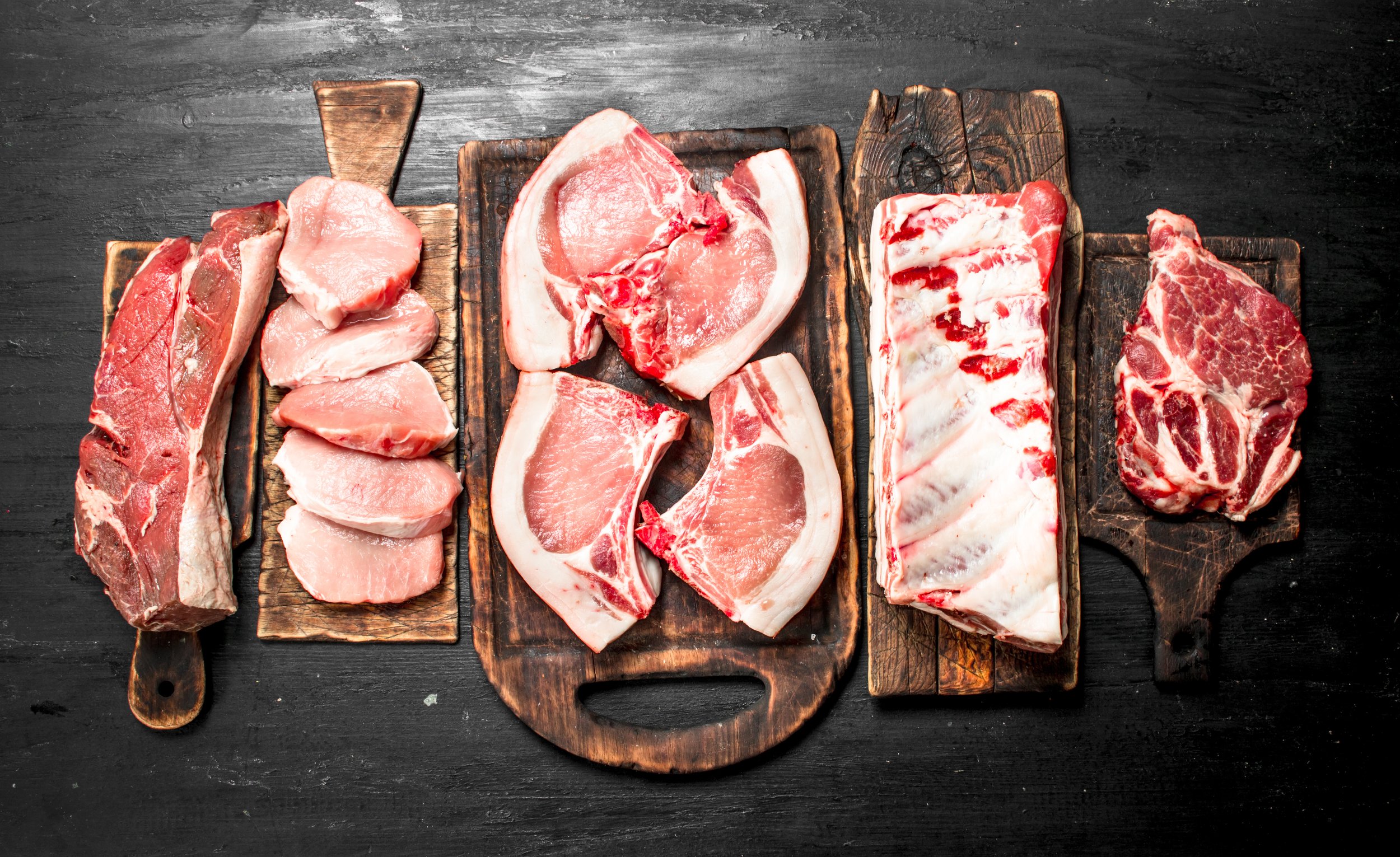 Assorted cuts of fresh meat displayed on wooden cutting boards, including pork chops, pork loin, ribs, and beef steak, arranged on a dark wooden surface. Ideal for meat lovers and culinary enthusiasts.