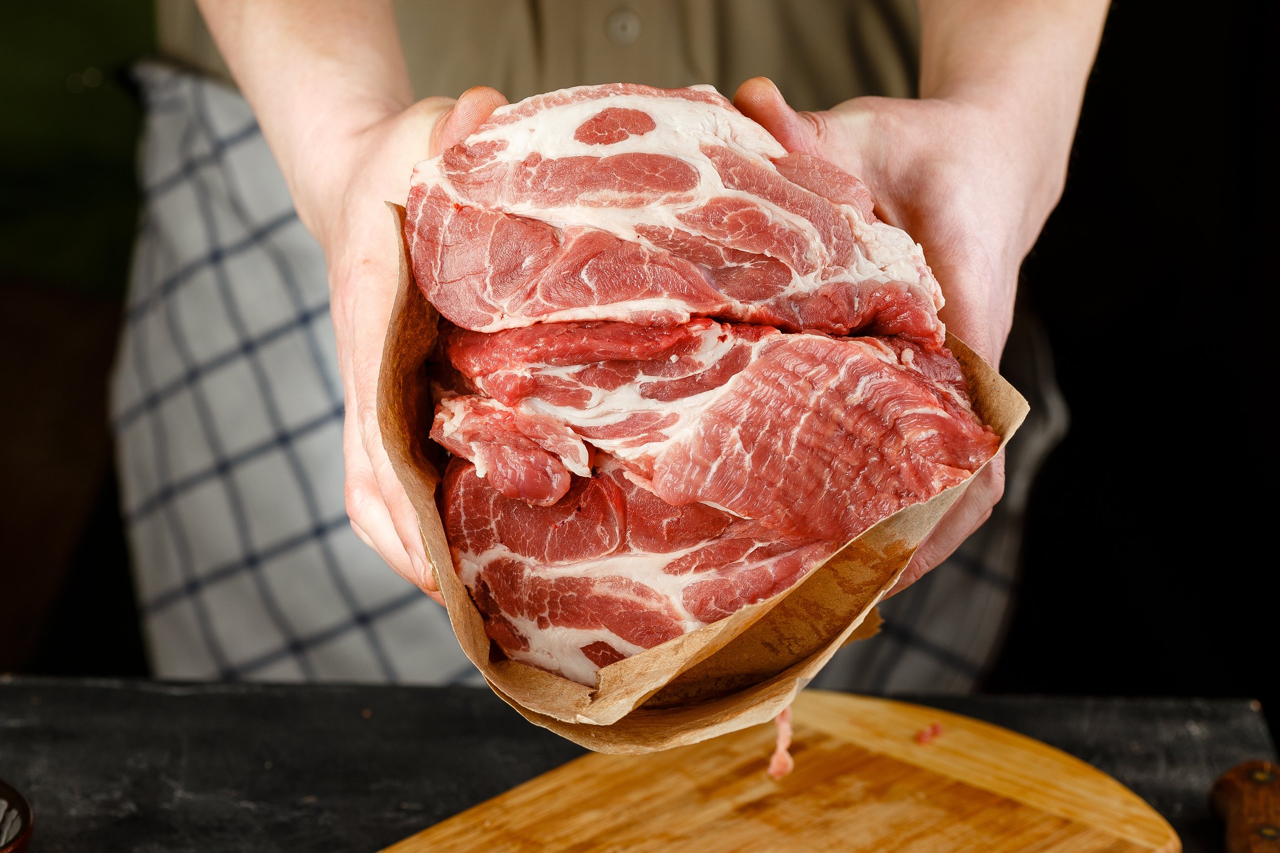 Fresh cuts of pork meat held in parchment paper, showcasing marbling and quality, with a wooden cutting board in the background. Ideal for cooking enthusiasts and meat lovers.
