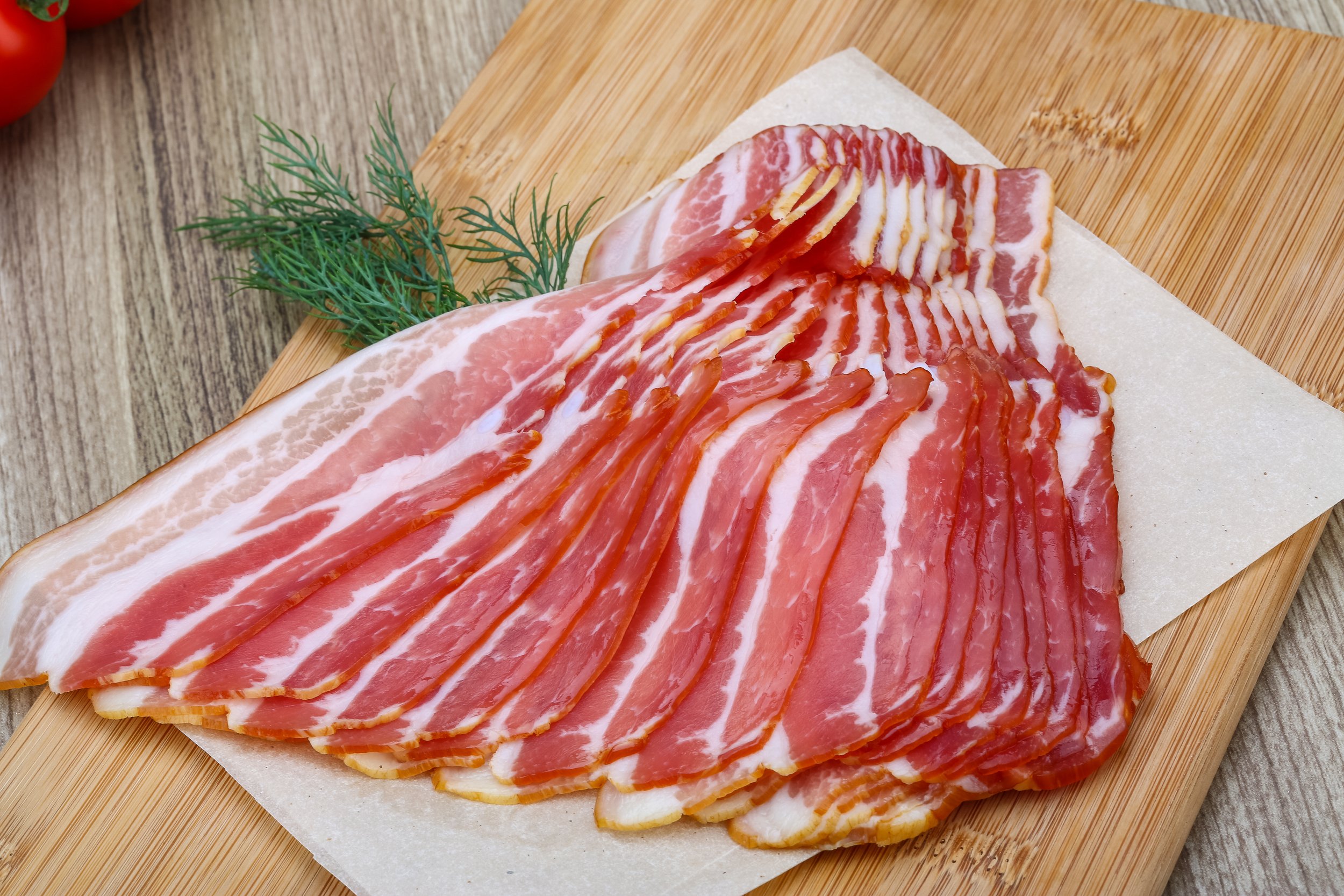 Sliced bacon arranged neatly on a wooden cutting board, with fresh dill and tomatoes in the background, showcasing the rich color and texture of the meat.