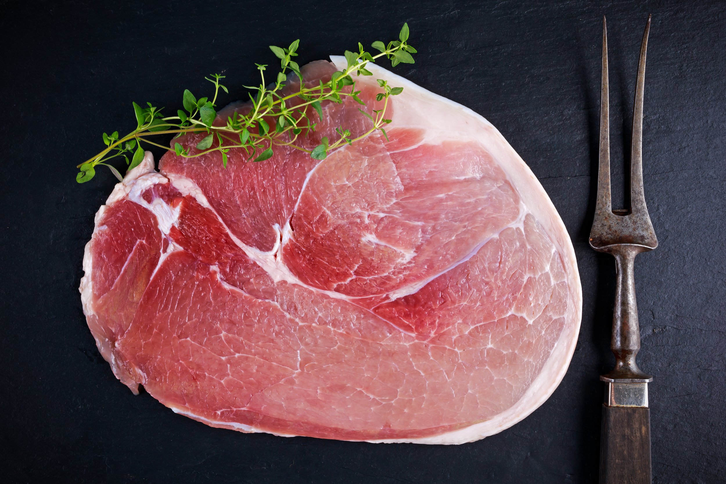 Raw pork chop with marbling, garnished with fresh thyme, and accompanied by a vintage meat fork on a dark slate background.