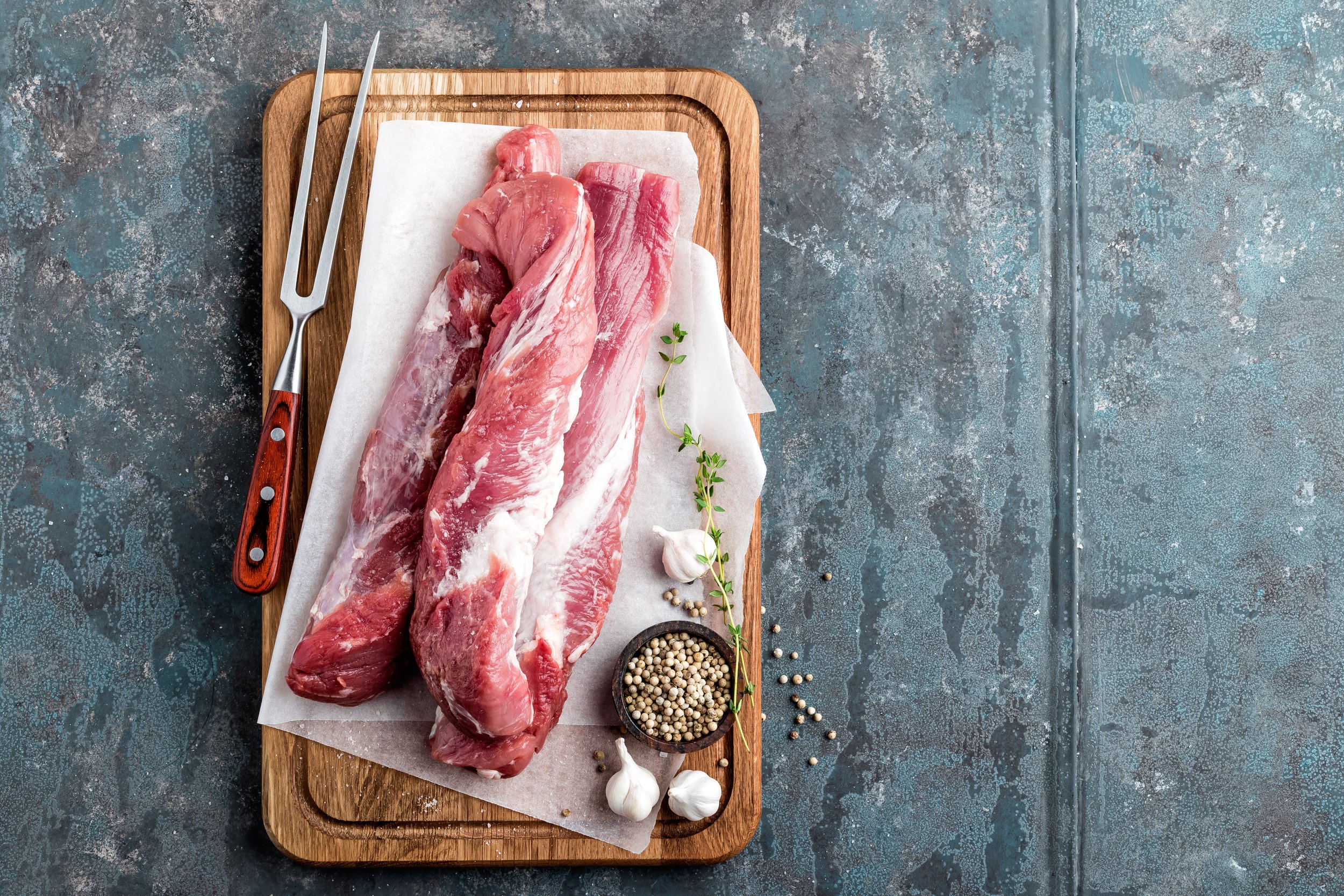 Fresh pork tenderloin displayed on a wooden cutting board with a pair of carving forks, accompanied by garlic cloves, green thyme, and a bowl of peppercorns, ideal for meal preparation and cooking inspiration.